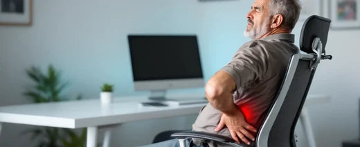 Homem sentado em uma cadeira ergonômica em um escritório, demonstrando desconforto nas costas, com a mão apoiada na região lombar