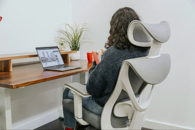Imagem de mulher sentada em cadeira ergonômica branca. A garota segura um caderno e assiste um vídeo em um notebook.