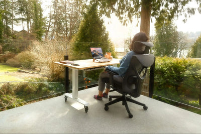 Imagem de mulher sentada em uma cadeira ergonômica preta, à sua frente há uma mesa de escritório com um notebook apoiado. A moça está em um mezanino e à sua volta há muitas árvores e plantas.