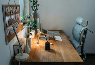 Imagem de escritório com janela e paredes brancas. No ambiente há uma cadeira ergonômica branca e mesa com notebook, luminária e vasos de planta.