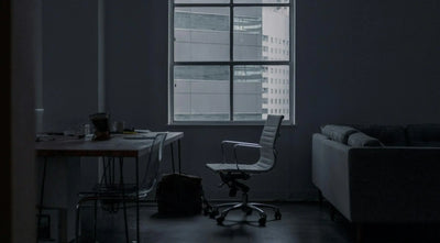 Imagem de escritório vazio com cadeira de escritório branca, uma mesa com objetos de trabalho, um sofá e uma janela grande com vista para prédios urbanos.